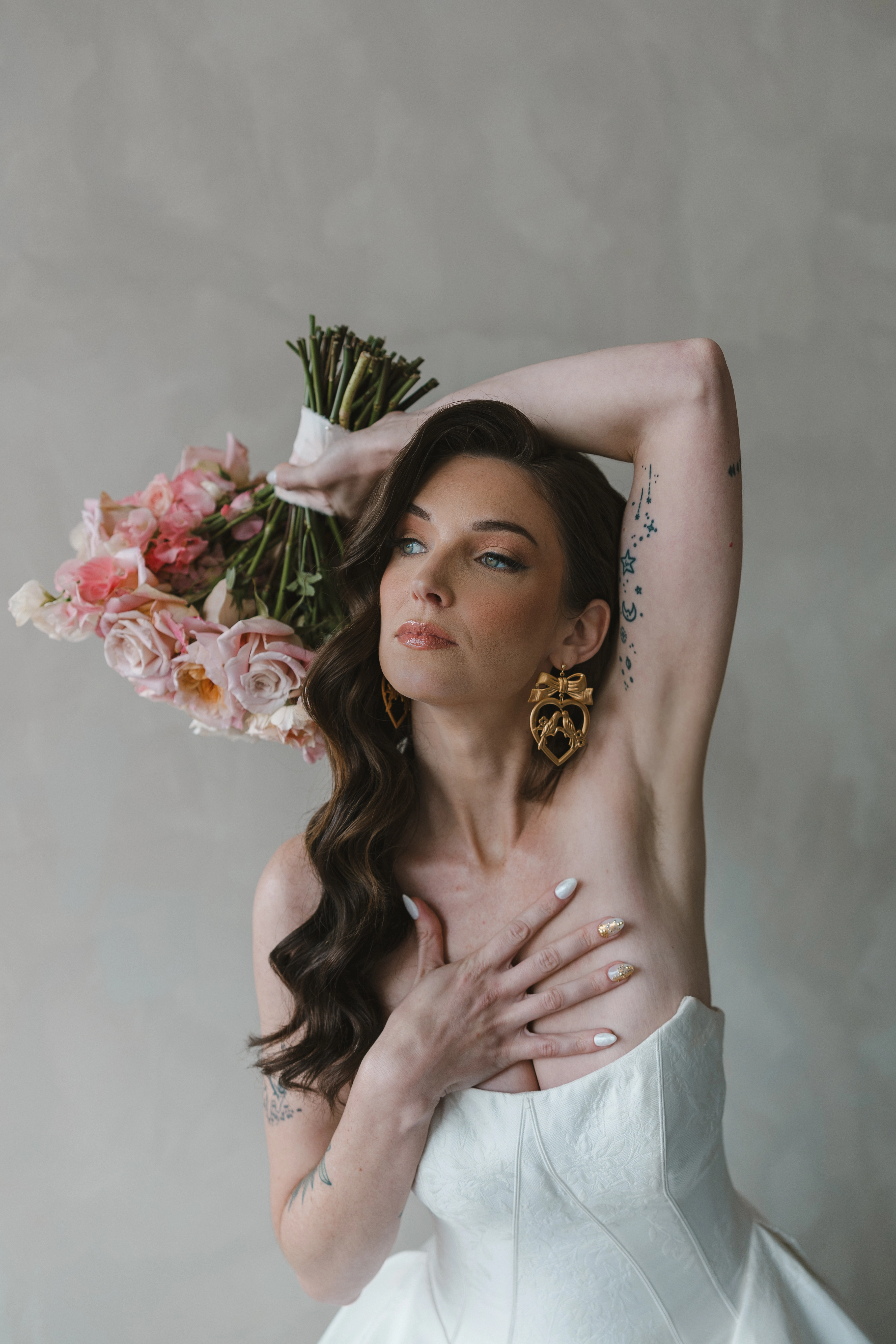 A bride wearing the 'Fortune Marie' strapless white dress by Maggie Sottero is posing with one arm raised and holding a bouquet of pink and white flowers. The bride's other hand is placed on their chest, and they are wearing gold earrings. The background is a plain, light-colored wall.